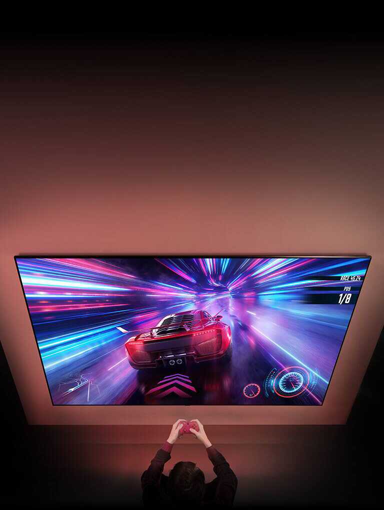 На стене закреплен большой телевизор, на экране которого видна гоночная игра. Перед телевизором вы можете видеть руки и контроллеры человека, сосредоточенного на игре.