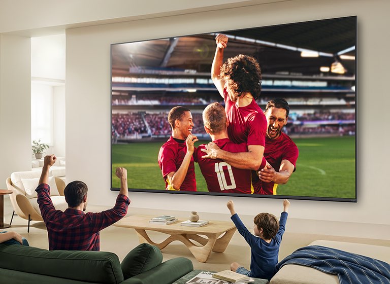 Ультрабольшой телевизор LG QNED evo AI Mini LED QNED8M показывает захватывающую крупномасштабную футбольную игру с улучшенным качеством изображения и яркими цветами, а семья вместе радуется на диване в гостиной.