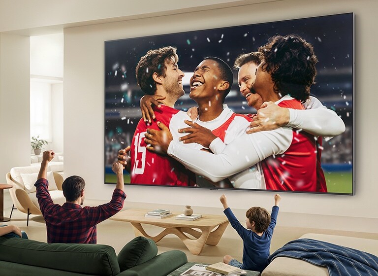 Телевизор LG QNED evo AI QNED90 Mini LED Ultra Big закреплен на стене в светлой гостиной, показывая большой триумф на стадионе во время спортивного события, с яркими цветами и великолепным качеством изображения на широком экране; сидящая на диване семья радуется.