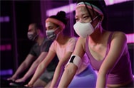 Женщина с носимым очистителем воздуха LG PuriCare™ катается на велосипеде по тренажерному залу, не вспотев. Рядом с ней мужчина и женщина в масках, кажется, едут на велосипедах с потом на лбу.