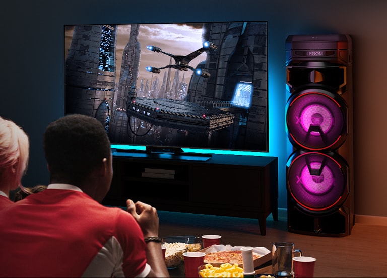 Люди смотрят телевизор на стене с системой LG XBOOM RNC9 справа от него. По телевизору показывают фильм. 