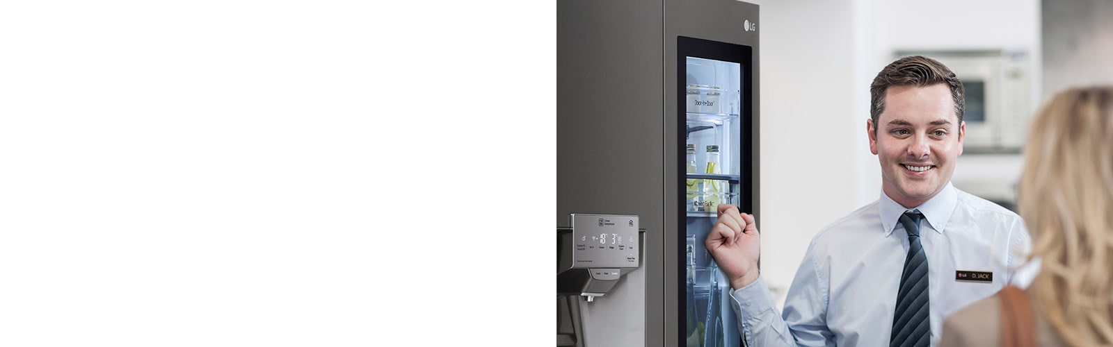Мужчина в рубашке и галстуке улыбается и разговаривает с женщиной, стоящей спиной к камере, указывая на холодильник позади себя.