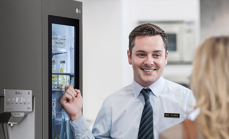 Мужчина в рубашке и галстуке улыбается и разговаривает с женщиной, стоящей спиной к камере, указывая на холодильник позади себя.