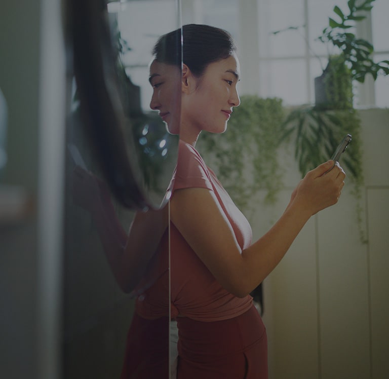 На изображении женщина стоит рядом со стиральной и сушильной машиной, смотрит в телефон и улыбается.