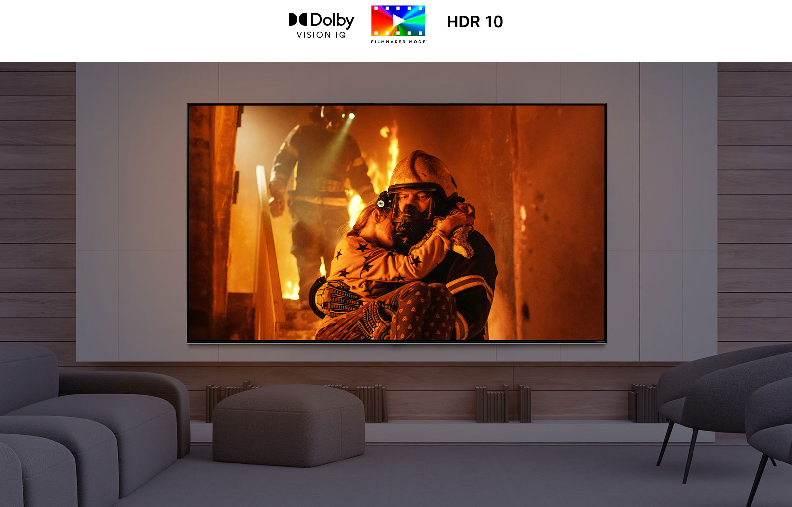 Телевизор с большим экраном, установленный на стене в комнате с несколькими низкими стульями. На экране показан мужчина, выносящий ребенка из пылающего здания.