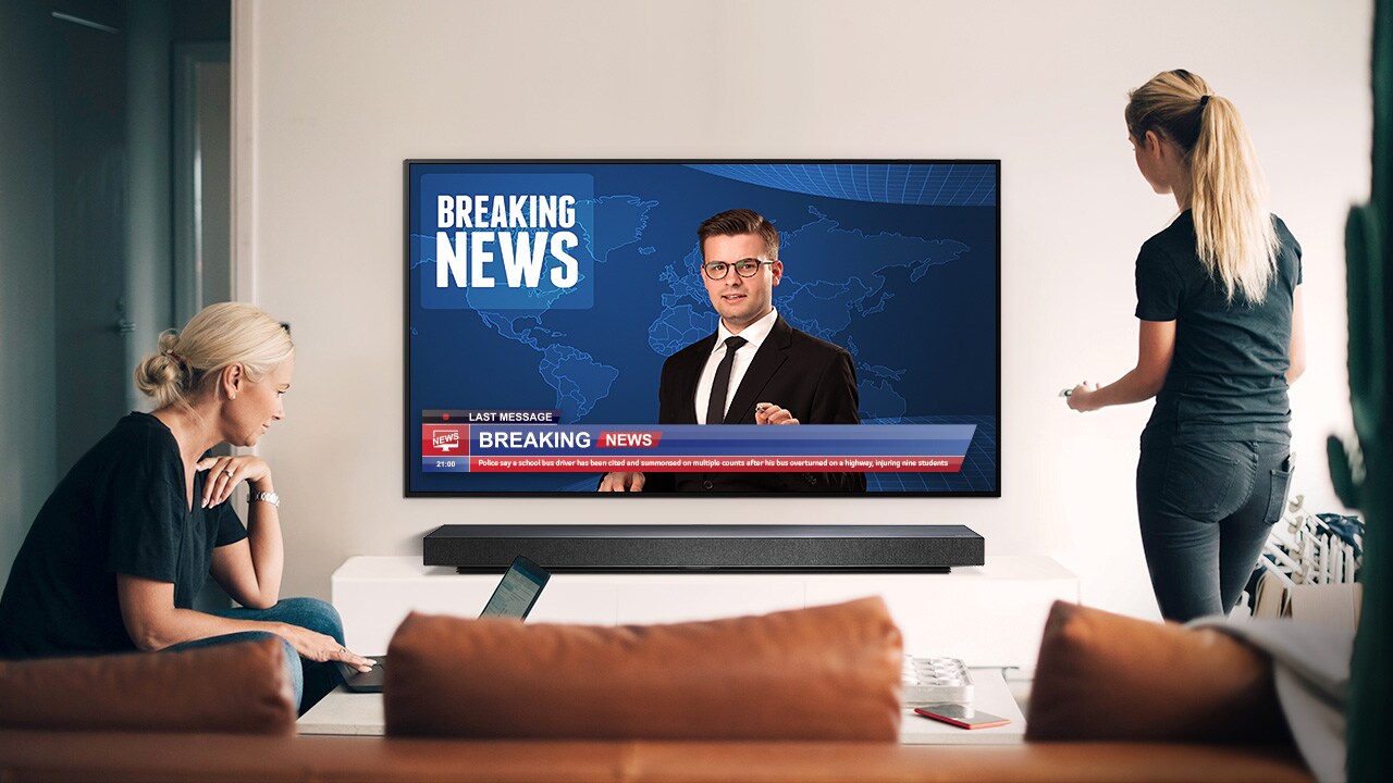 A woman watching the news at its best quality on TV standing up and another sitting in front of her laptop (move the slide)