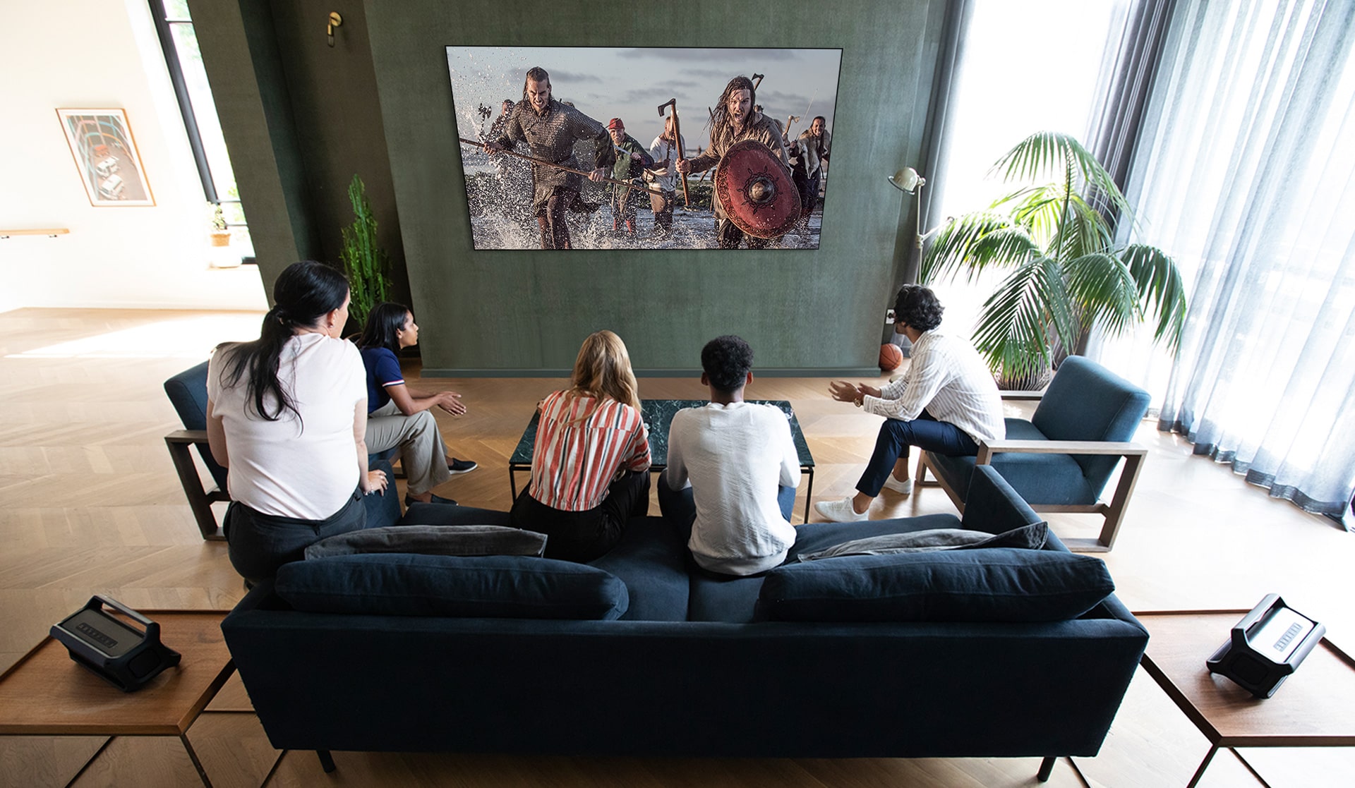 A group of friends watching a war movie on TV in the living room with 2 same Bluetooth speakers at rear speaker position