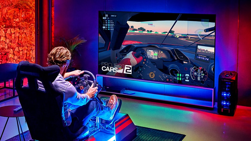 Man in a dark room, sitting on a racing game seat, in front of a large TV playing a racing game