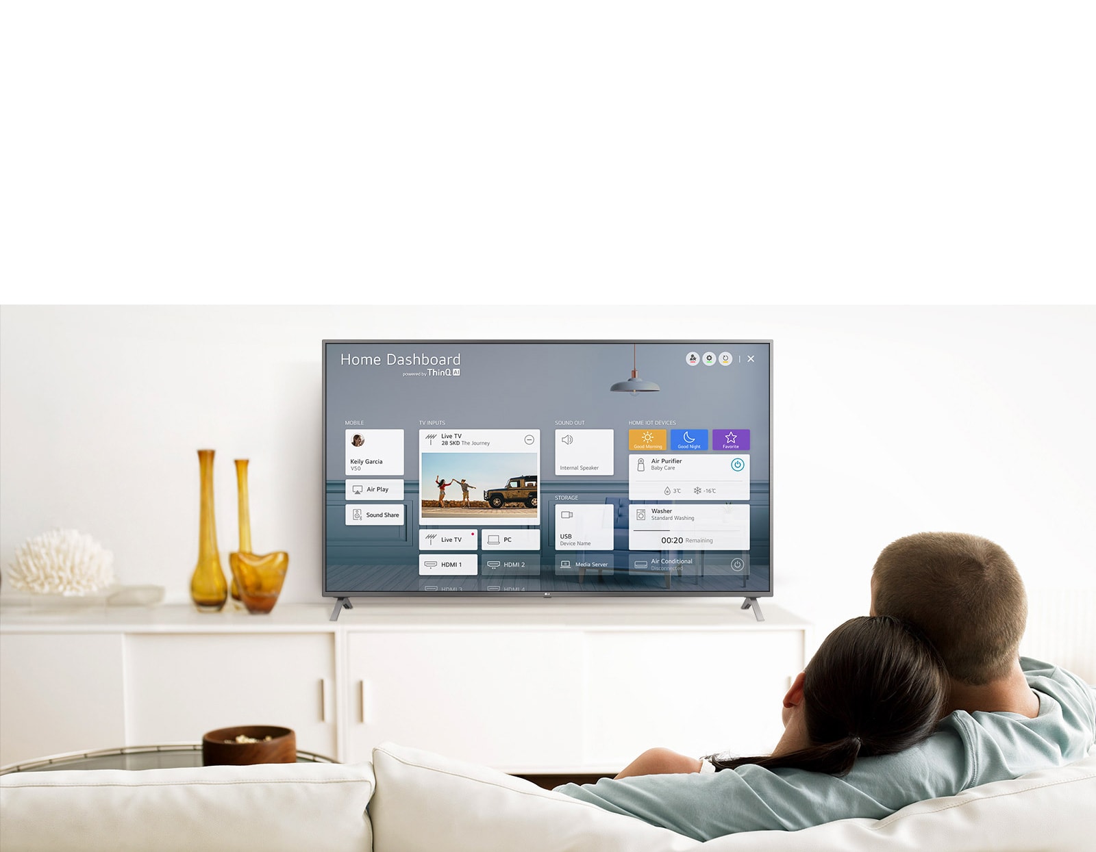 A men and women sitting on a sofa in the living room with the Home Dashboard on the TV screen