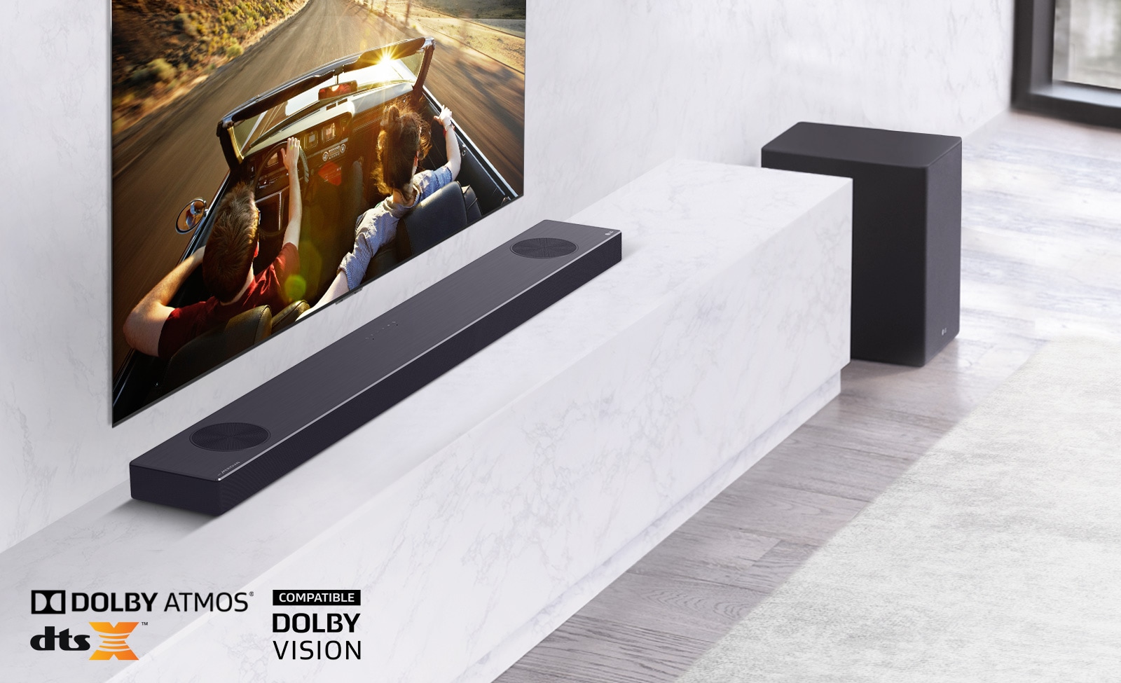 TV is on the wall, LG Soundbar is below on a white marble shelf with a sub-woofer to the right. TV shows a couple in a car. 