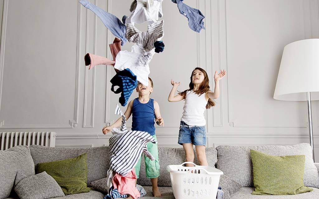 Two children throwing laundry from the basket on the sofa, in the living room with cushions and lamp in the background