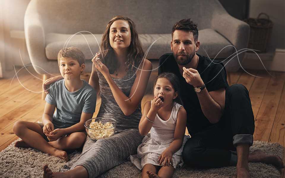A family eating popcorn whilst watching their OLED TV with sound waves displayed around them.