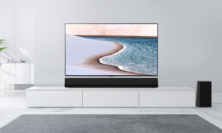 Televízor osadený na svetlosivej stene. LG Sound Bar je umiestnený pod ním na bielej skrinke. Na obrazovke televízora je pláž.