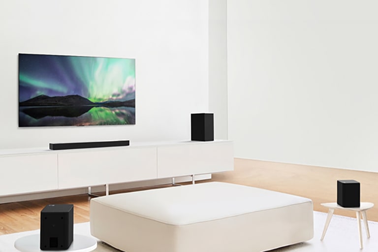 Televízor a Sound Bar v bielej obývačke s bielou pohovkou uprostred. Na krajoch pohovky sú položené reproduktory.