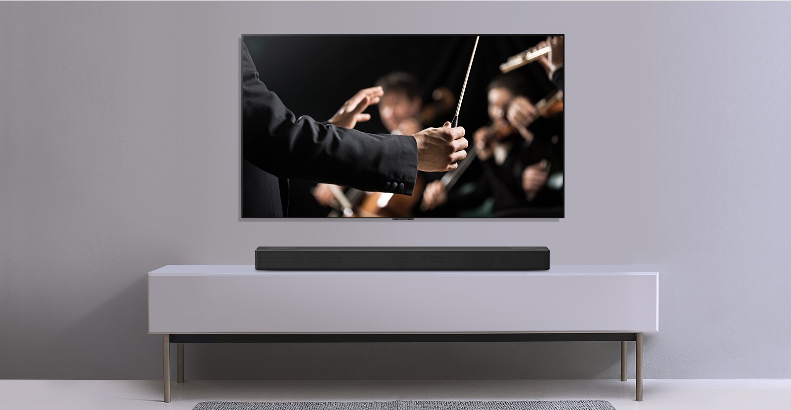 Televízor je umiestnený na sivej stene a pod ním je LG Sound Bar na sivej polici. Na obrazovke televízora je dirigent, ktorý diriguje orchester.