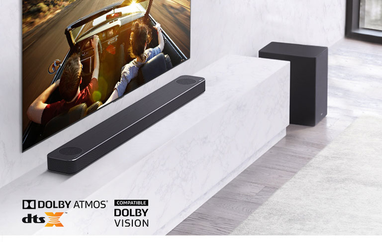 Televízor je umiestnený na stene, pod ním sa nachádza LG Sound Bar na mramorovej bielej polici so subwooferom na pravej strane. Na obrazovke televízora je pár v aute.
