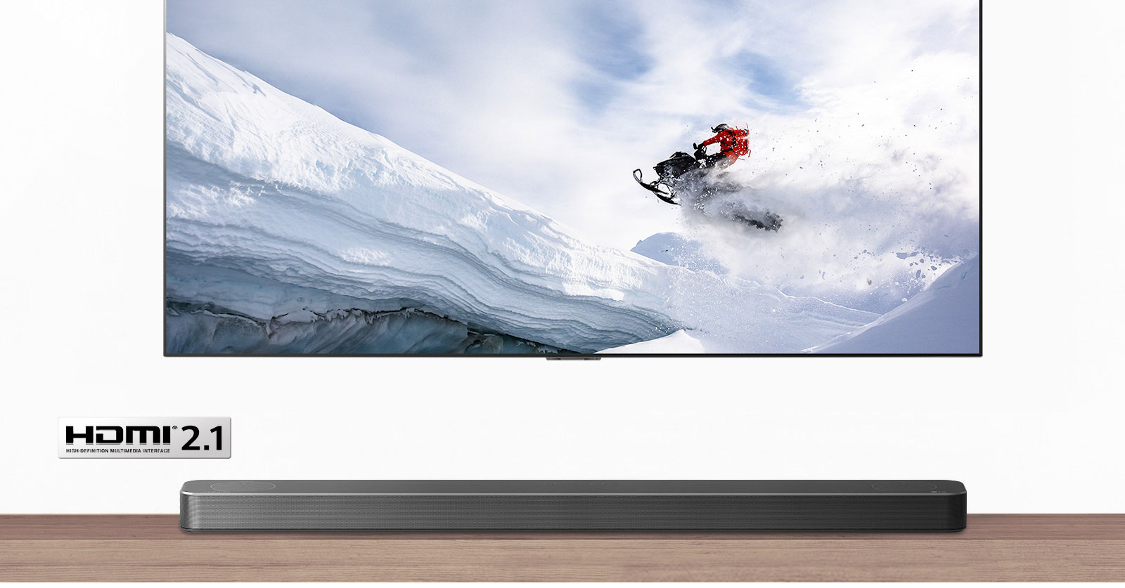Televízor a soundbar v pohľade spredu. V televízore ide muž na snežnom skútry zasneženými horami. Logo HDMI 2.1 je pod televízorom.