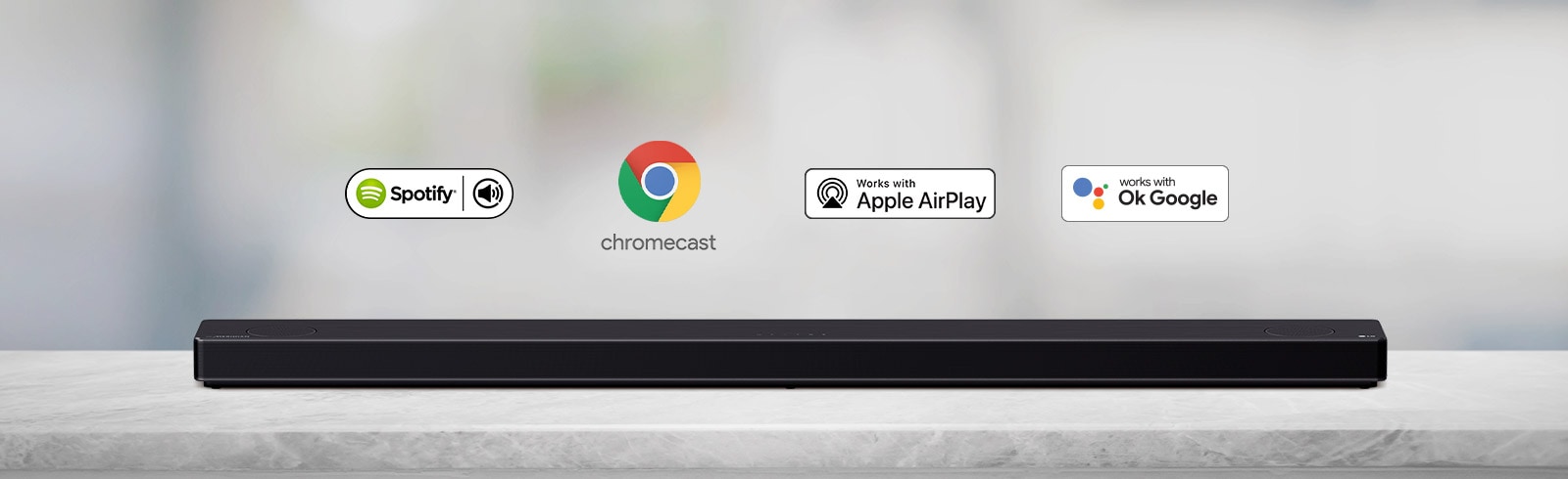 Na sivej polici je položený zvukový panel. Zľava doprava sú zobrazené logá platforiem AI v poradí: Spotify, Chromecast, Apple Airplay a OK Google.