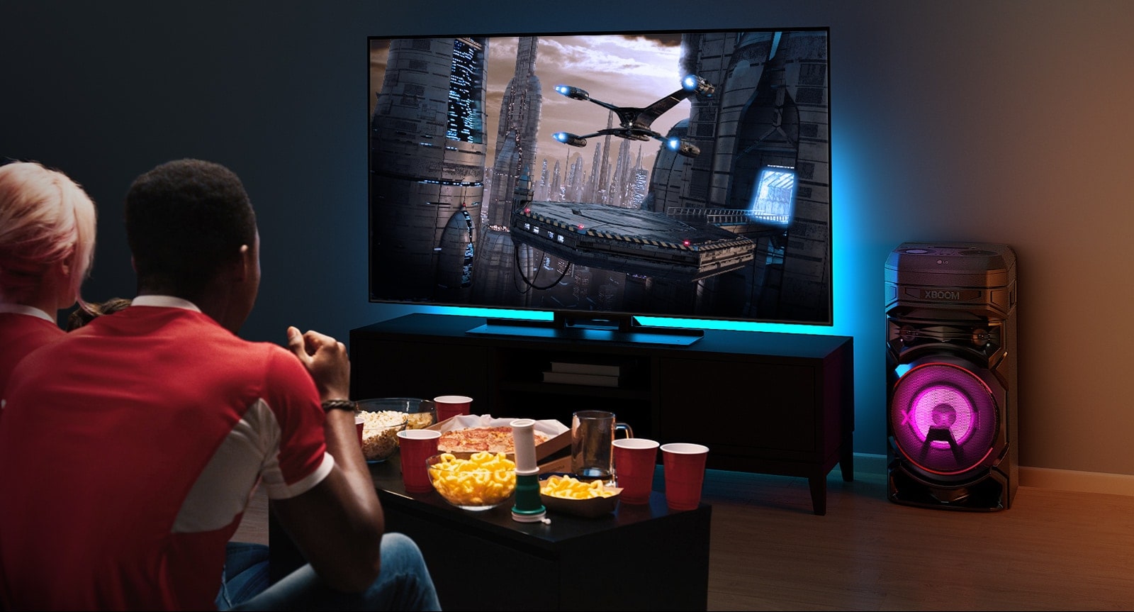 Ľudia sledujú televízor na stene s reproduktorom LG XBOOM RNC5 napravo od neho. Na televízore sa prehráva film. 