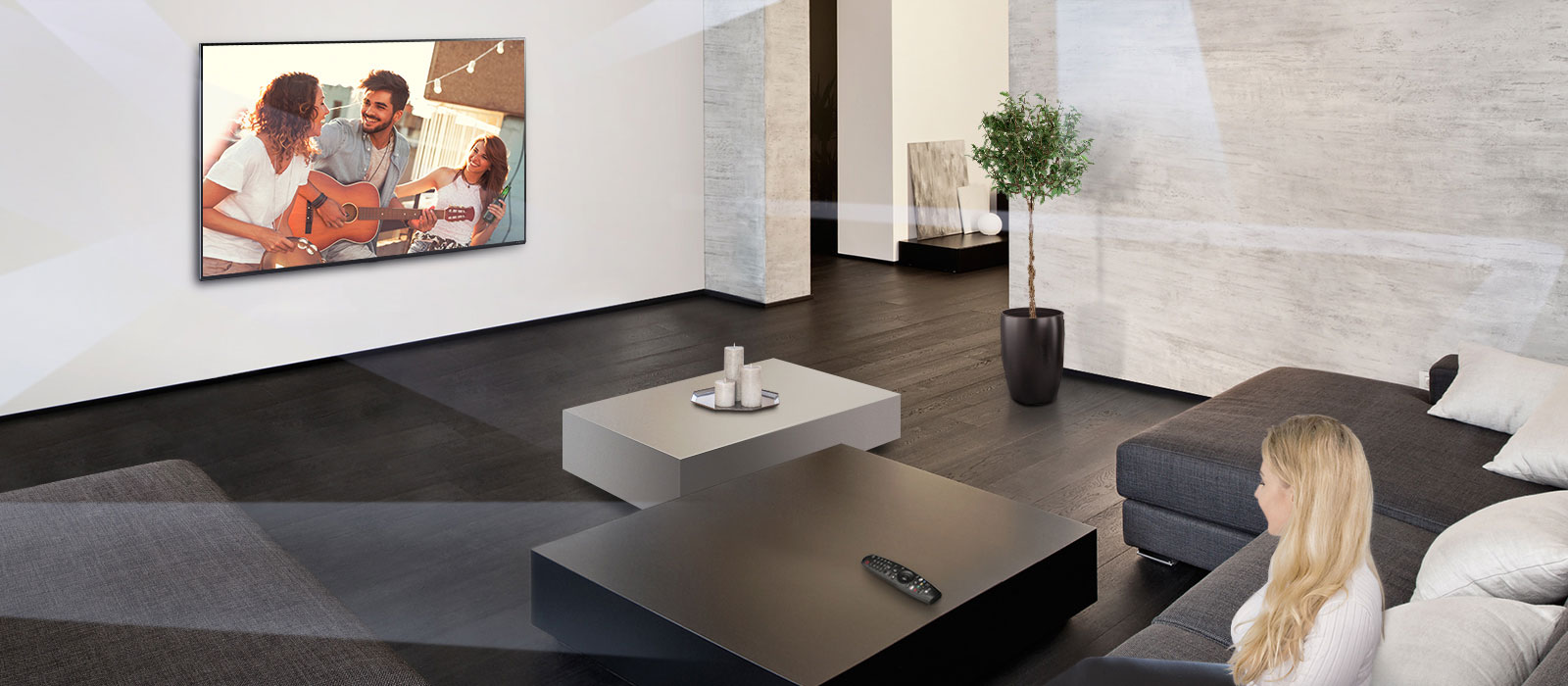 Široký záber na presvetlenú miestnosť s televízorom osadeným na stene a ženou sediacou na gauči oproti nemu a sledujúcou televízor.