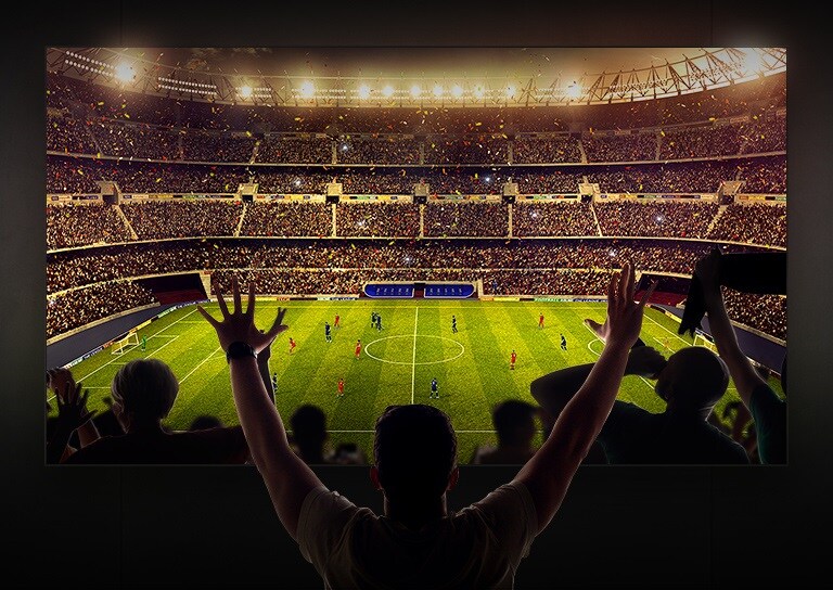 Štyria ľudia sedia na pohovke a sledujú televízor s veľkou obrazovkou, na ktorom je zobrazený futbalový štadión plný divákov.