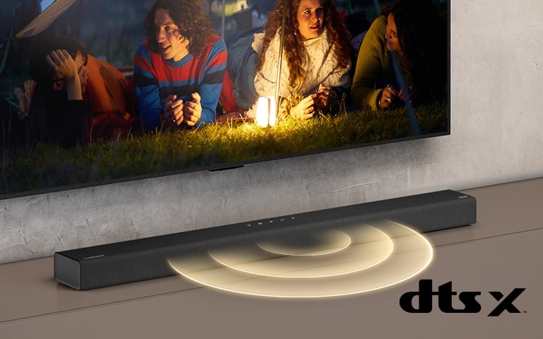 LG TV je na stene, na obrazovke vidno dva páry ležiace na tráve. Pred nimi je lampa. Zvukový panel LG sa nachádza pod televízorom LG. Z prednej časti zvukového panela vychádza zvuková grafika. Logo DTS Virtual:X je zobrazené v pravom dolnom rohu obrázka.
