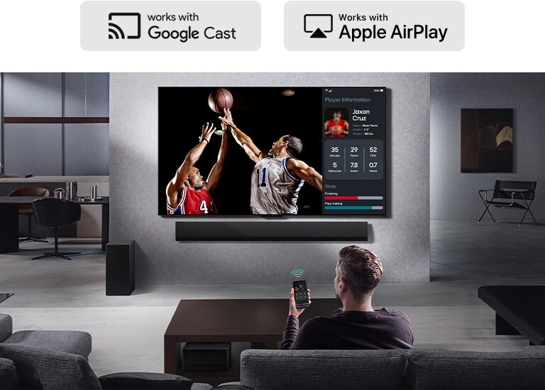Une personne dans son salon qui tient son téléphone. Sur le téléphone se trouve une icône de transmission montrant que l’écran du téléphone est mis en miroir sur la TV. Sur la TV, on voit un match de basket et sur le côté se trouve l’écran mis en miroir, montrant les statistiques du joueur. 