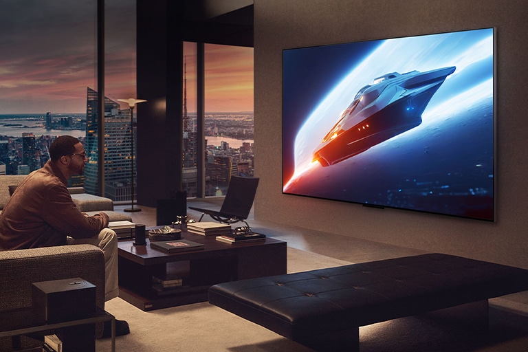 Salon élégant dans un appartement penthouse. Magnifique paysage urbain visible à l’extérieur de la fenêtre. Un homme assis sur le canapé regarde du contenu sur une TV fixée au mur. 