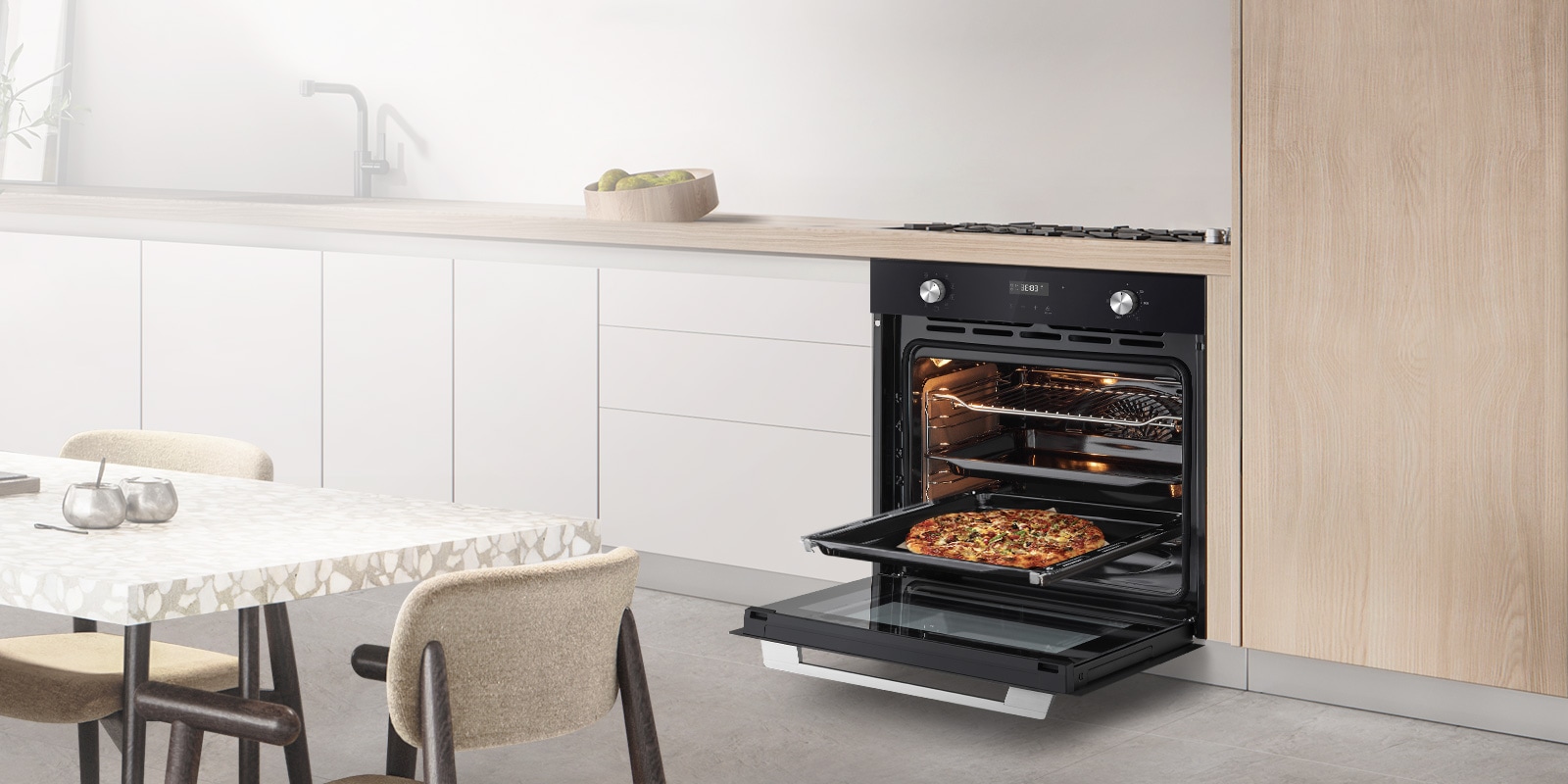 Вбудована духова шафа встановлена на сучасній білій кухні. Духова шафа відкрита, на нижньому деко готується піца.