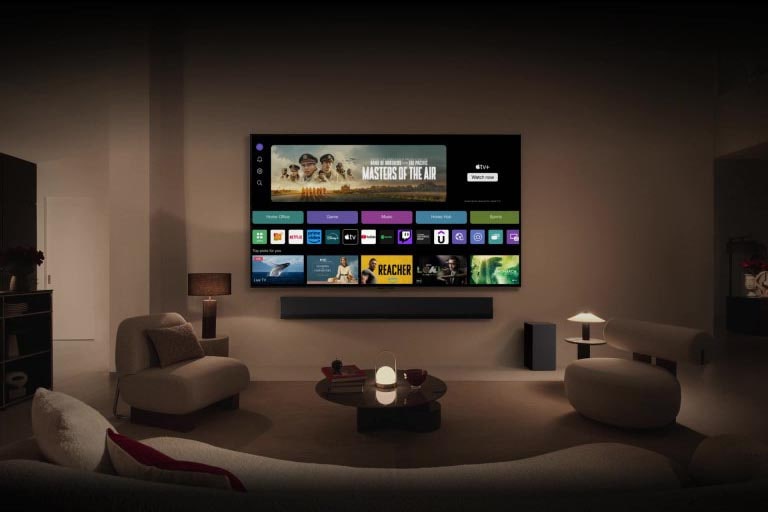 A close-up of an LG TV screen showing the buttons Home Office, Game, and Music over a banner for Masters of the Air zooms out to show the TV mounted on a wall in a living room. The following logos are displayed on the TV screen in the image: LG Channels, Netflix, Prime Video, Disney TV, Apple TV, YouTube, Spotify, Twitch, GeForce Now and Udemy.