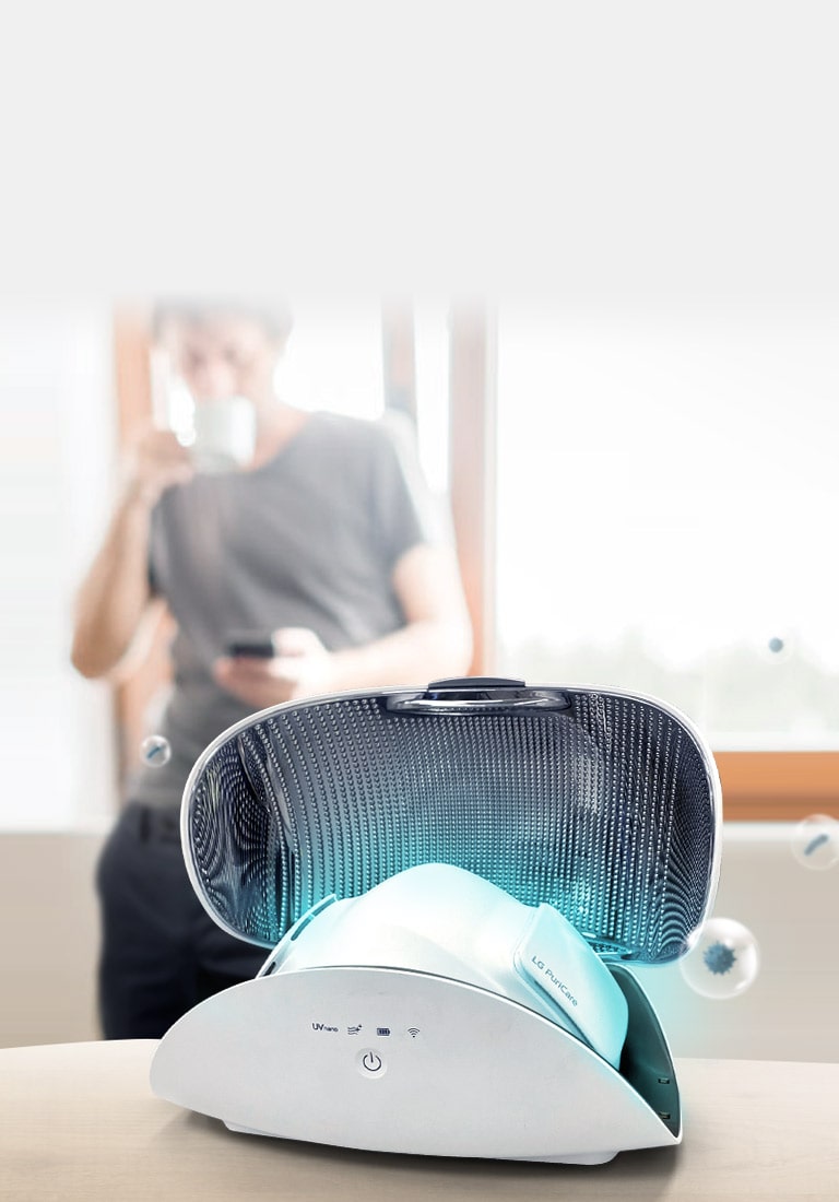 Очисник повітря індивідуального використання LG PuriCare™ показано всередині кейсу для нього на столі на передньому плані. Слабке блакитне світло відходить від маски, а бульбашки, що зображають шкідливі забрюднювачі, зникають вдалині. Чоловік п'є каву на фоні. 