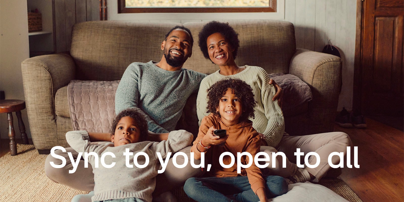 Зображення сім’ї із чотирьох осіб, які сидять на підлозі перед затишним коричневим диваном, дивляться телевізор і сміються. Поверх зображення напис білим шрифтом: «Синхронізовано з вами, відкрито для всіх». 