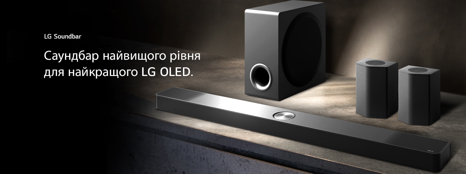 LG Soundbar, задні динаміки та сабвуфер (вигляд під кутом) на коричневій дерев’яній полиці в чорній кімнаті, огорнутій темрявою, де світло падає лише на звукову систему.