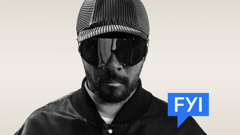 Black and white photo of will.i.am wearing a cap and sunglasses and facing the camera, with the FYI RAiDiO logo in the bottom right corner.