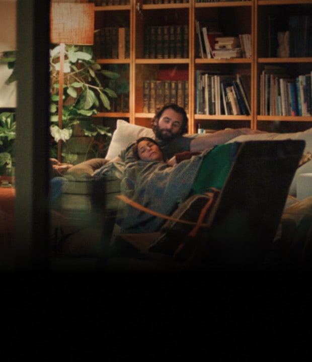 A scene where the camera captures a man and a woman lying together on the living room sofa from a distance.