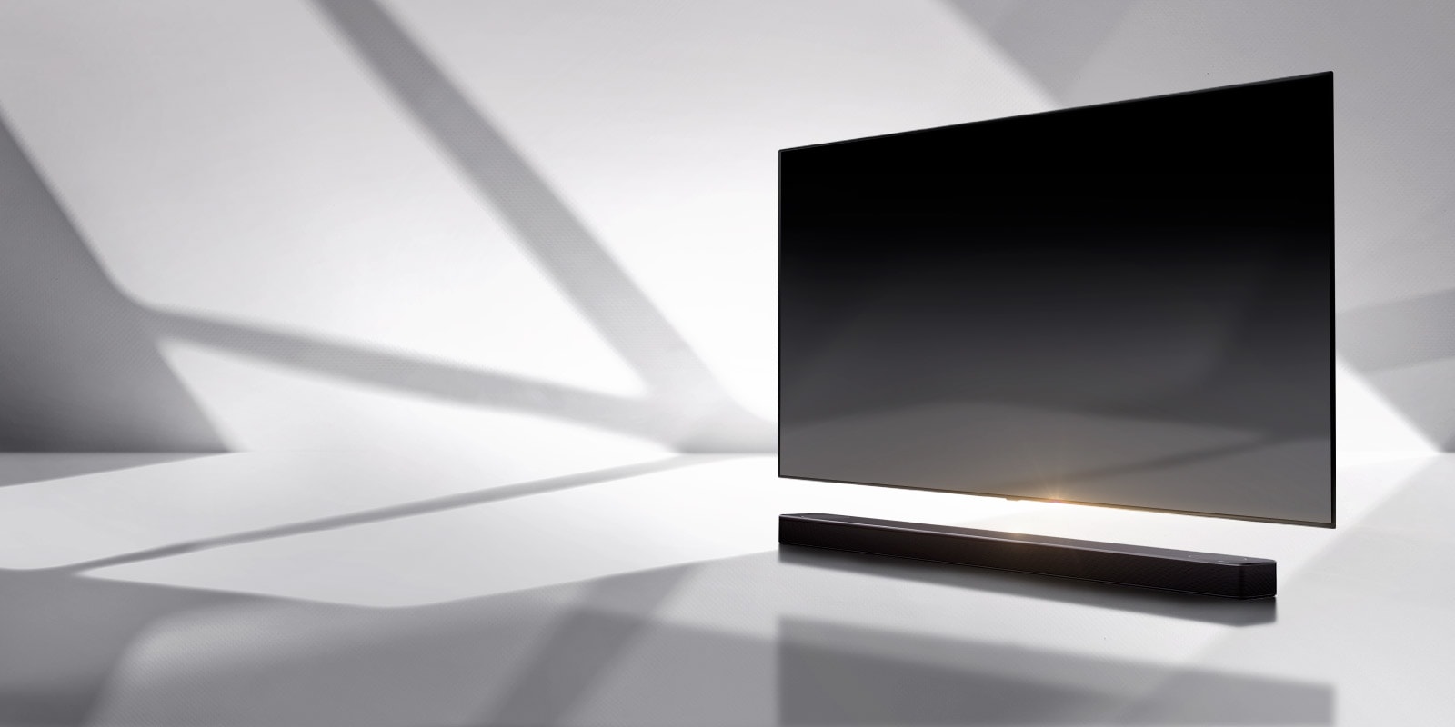 A soundbar and a TV are placed on a white floor and there is a shadow coming from outside right behind.