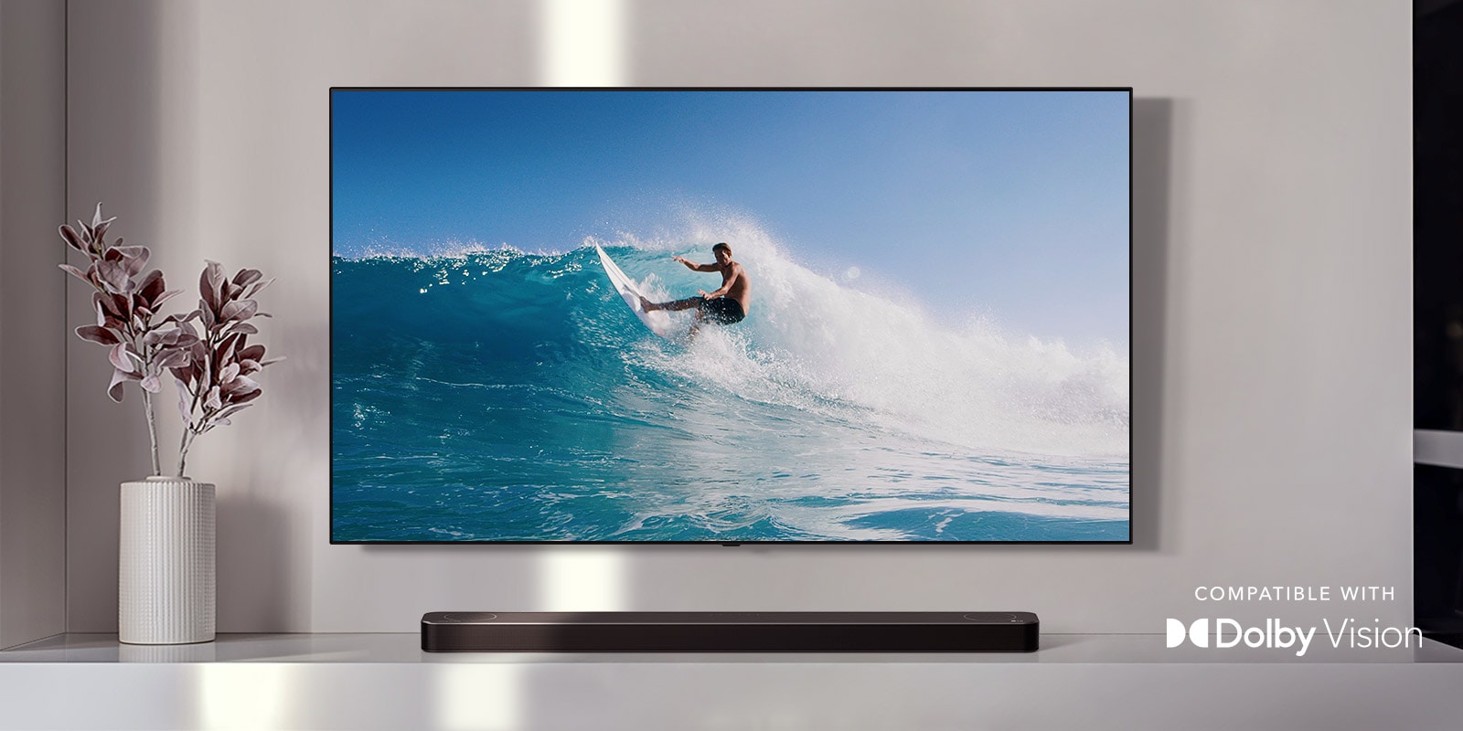 TV is on the wall. TV shows a man surfing on big wave. LG Soundbar is right below TV on a white shelf. There is a vase with a flower right next to the soundbar. (play the video) 