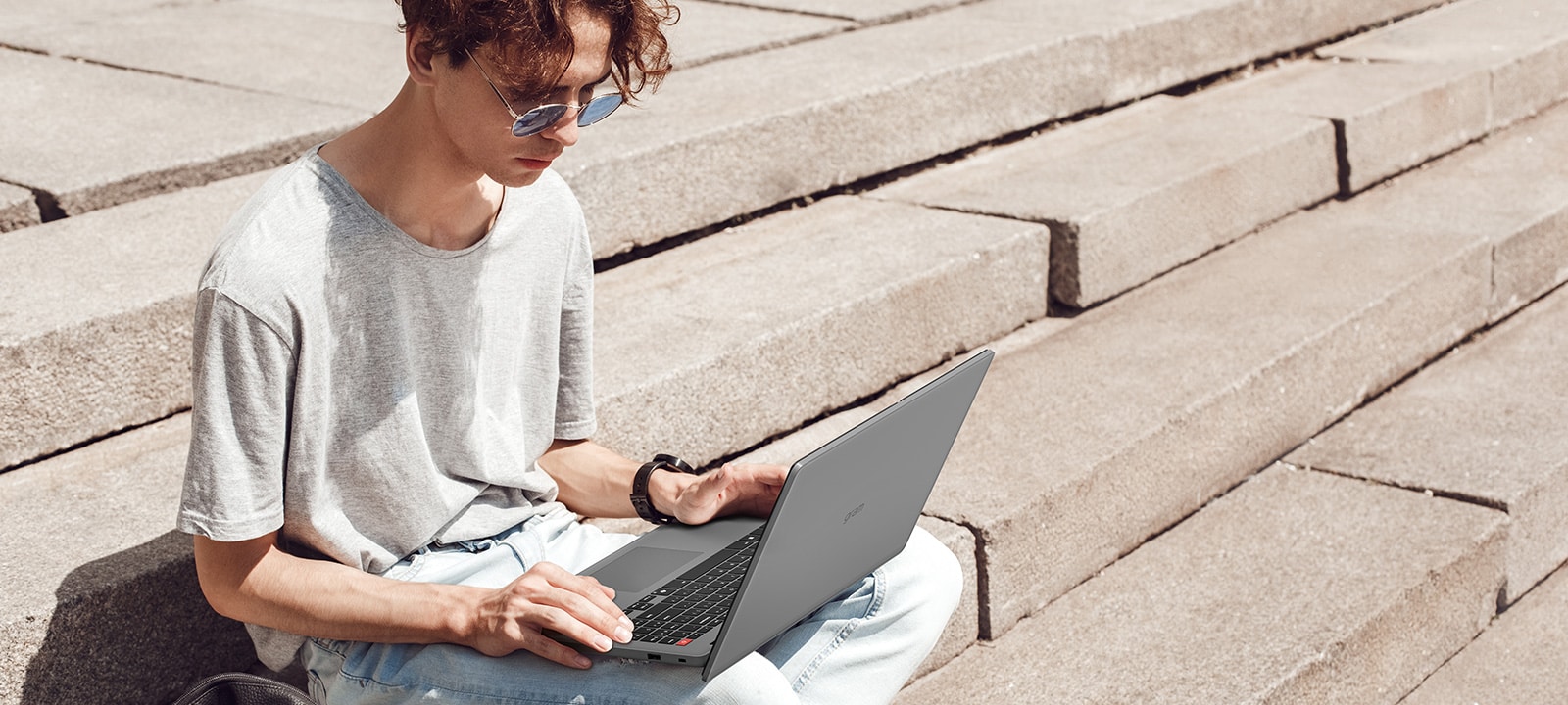 The image shows a young man sitting on outdoor concrete steps, using a gray laptop.The man appears focused on his work or browsing, highlighting the laptop's portability and suitability for on-the-go use.