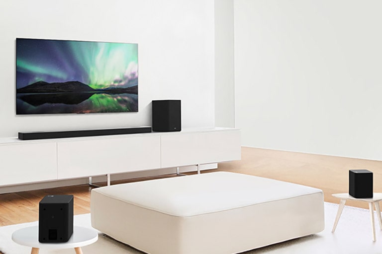 TV and sound bar in white living room with white sofa in the center. Speakers sit on both ends of the sofa.