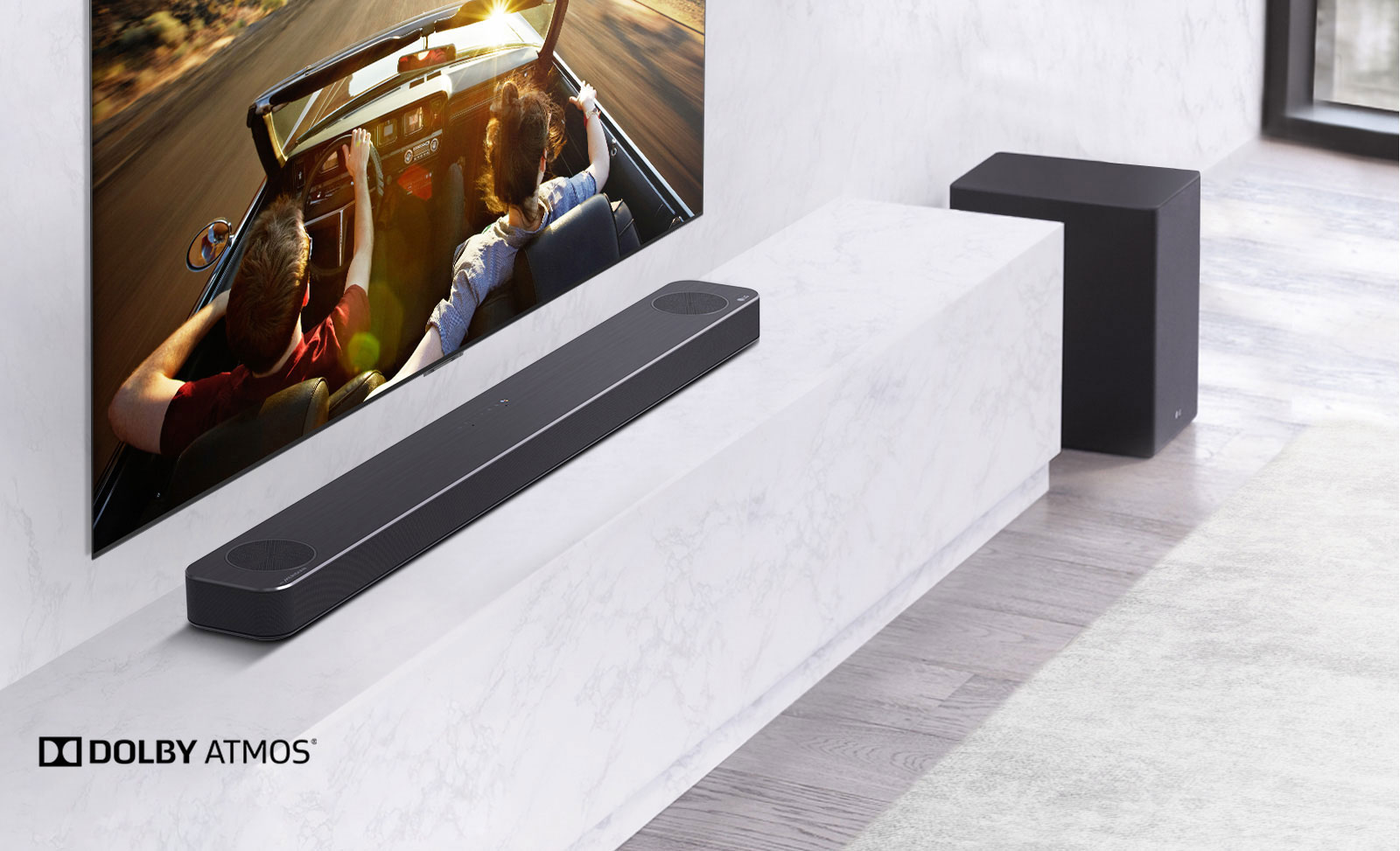 TV is on the wall, LG Soundbar is below on a white marble shelf with a sub-woofer to the right. TV shows a couple in a car.