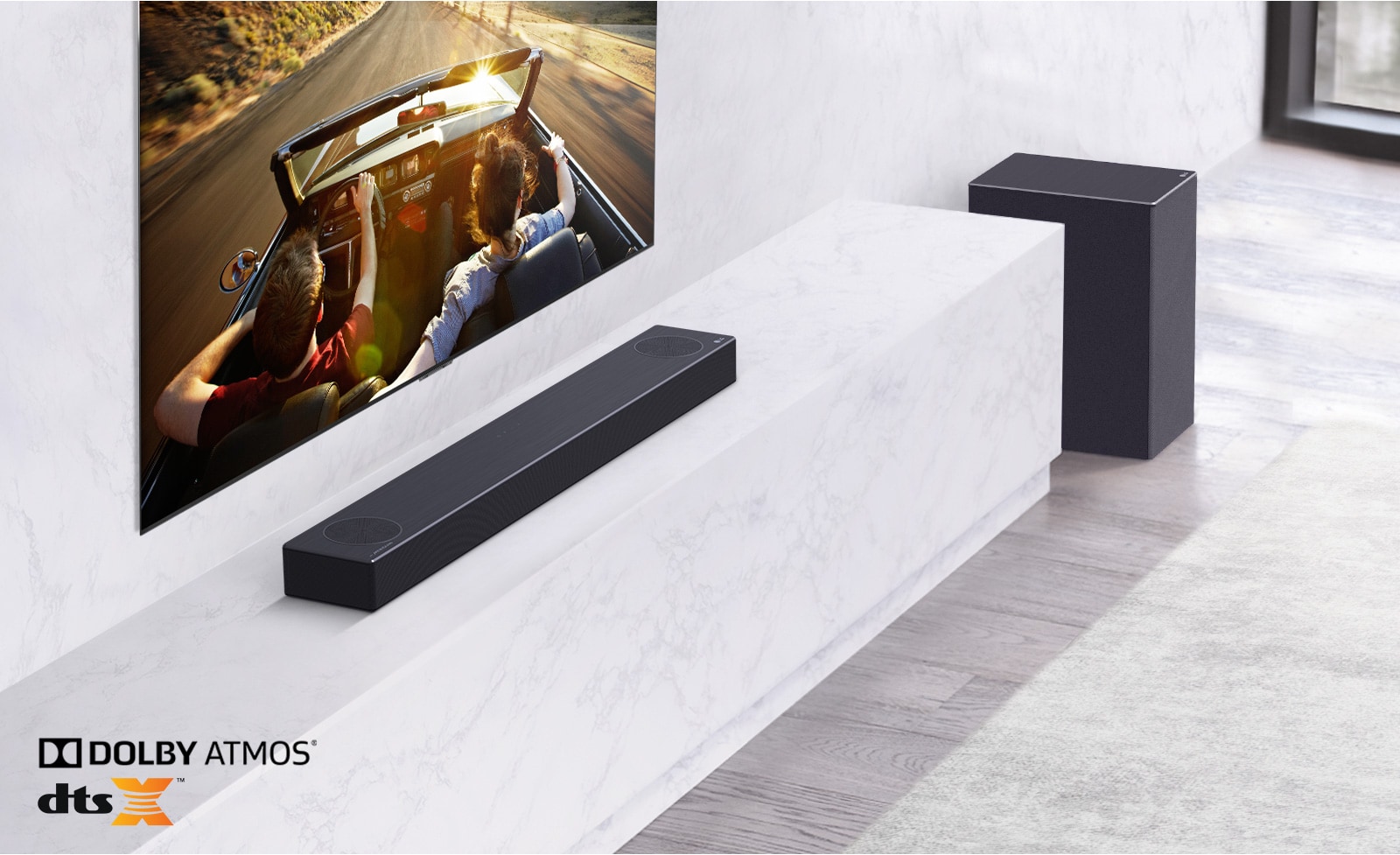 TV is on the wall, LG Soundbar is below on a white marble shelf with a sub-woofer to the right. TV shows a couple in a car.