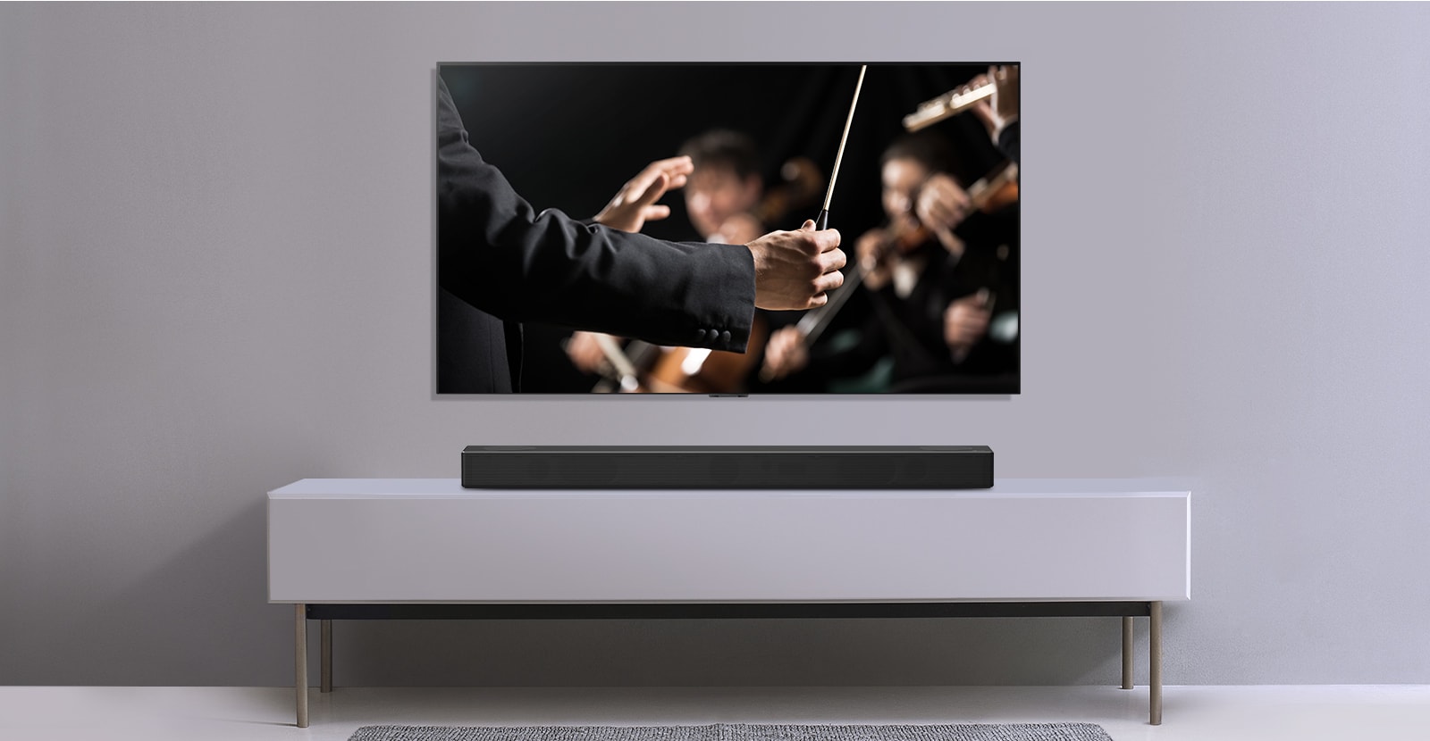 A TV is shown on a gray wall and LG Soundbar below it on a gray shelf. TV shows a conductor conducting an orchestra. 