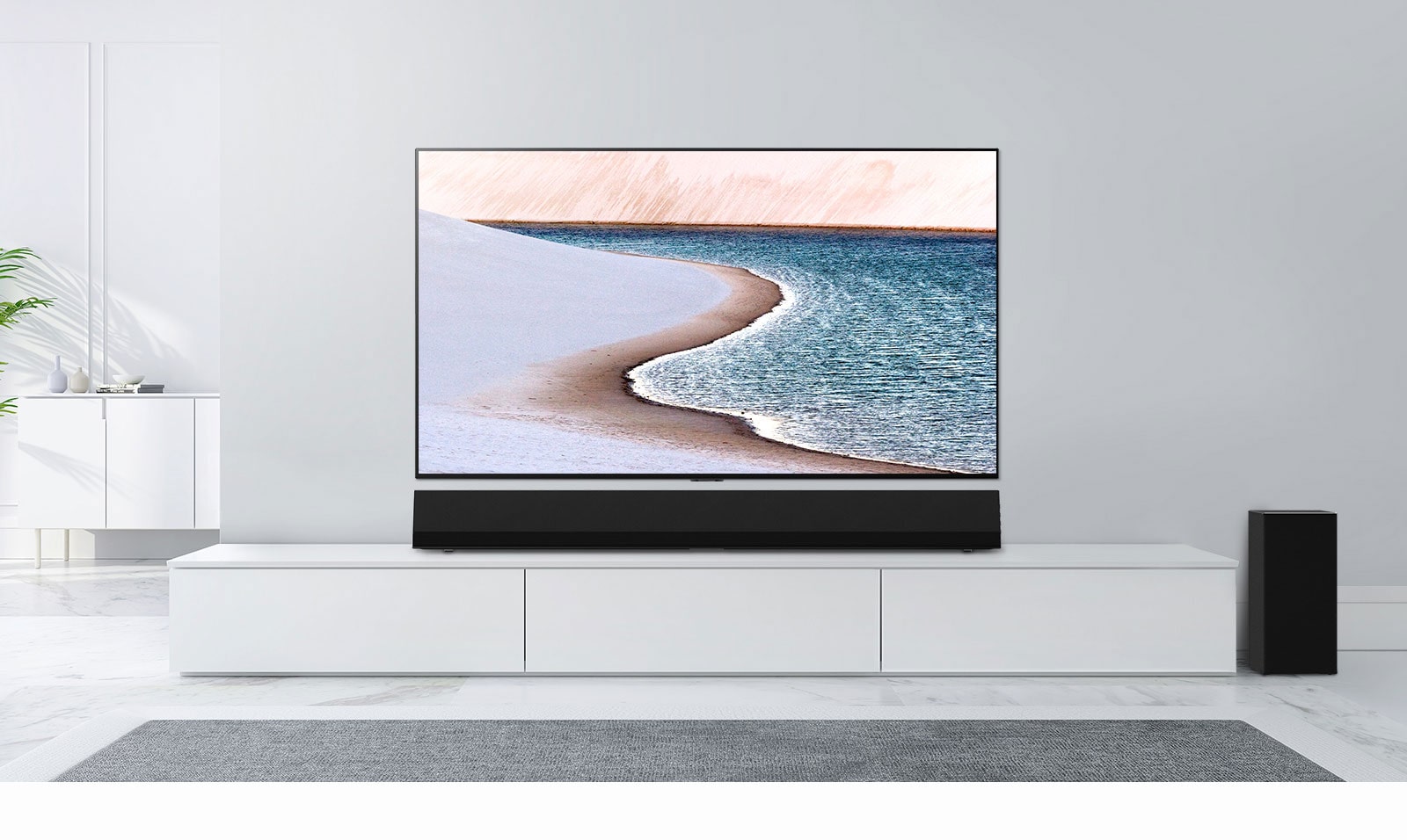 Na svetlo sivi steni je nameščen televizor.  LG Soundbar je pod njim na beli omari.  Televizor prikazuje plažo.