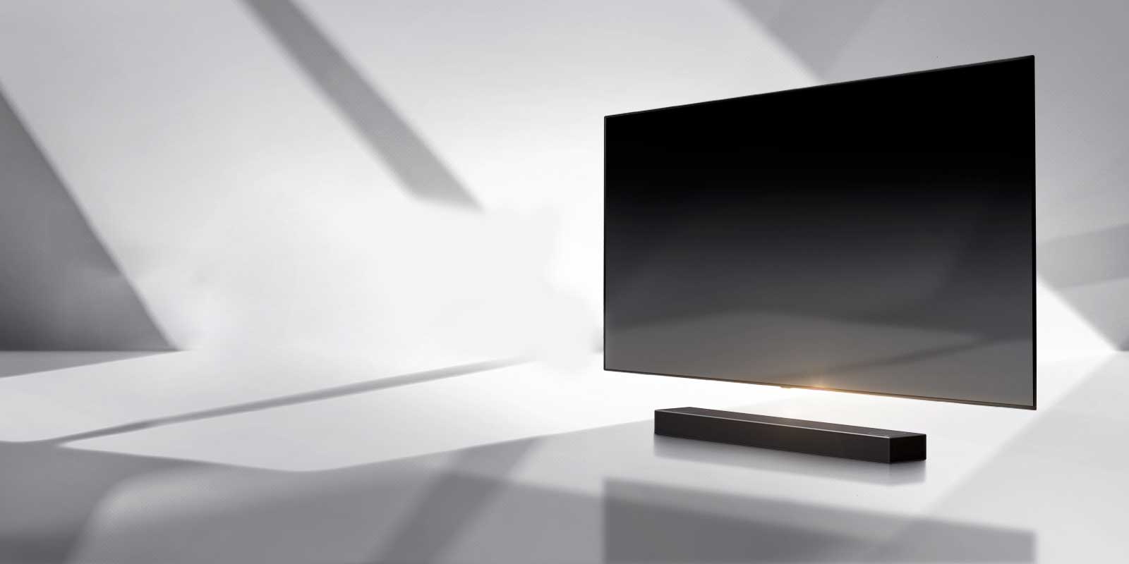 A soundbar and a TV are placed on a white floor and there is a shadow coming from outside right behind.