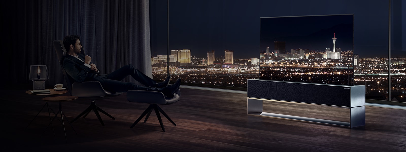 A man watches LG SIGNATURE's Rollable OLED TV facing the night city view of Las Vegas in his living room.
