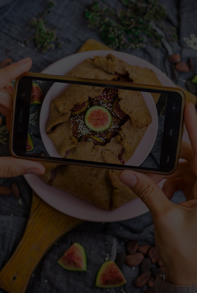 A pair of hands hold a smartphone over a fig pastry while snapping a picture. 'We are what we eat' is superimposed over it.