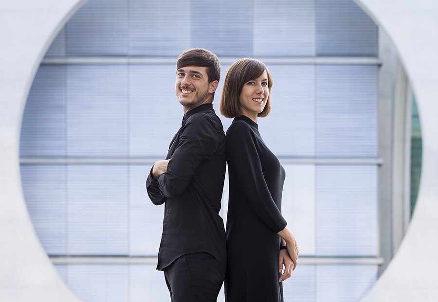 creator anna devis and daniel rueda are standing back to back and smiling