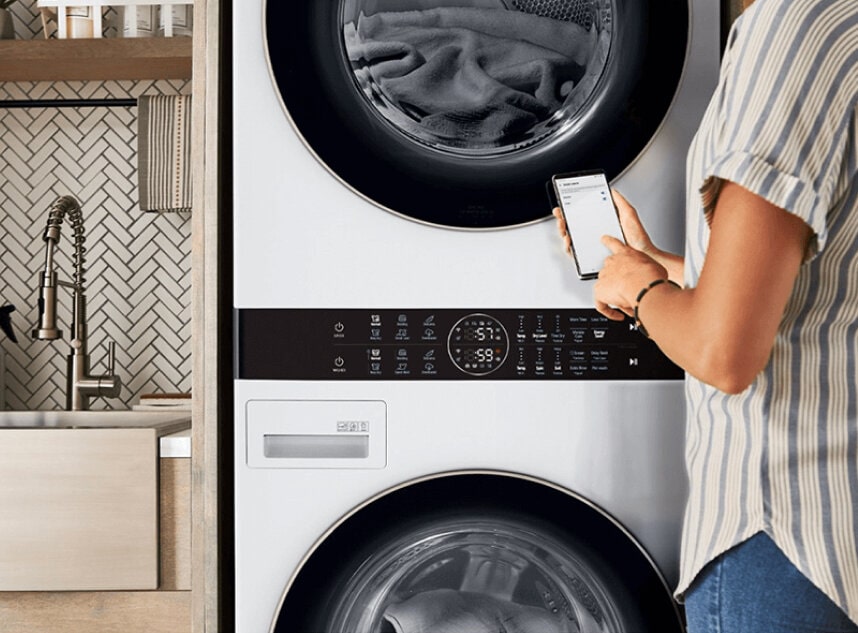 A person using the LG ThinQ® app on a smartphone in front of an LG Studio washer, emphasizing the reliability and 2-year warranty of LG appliances.