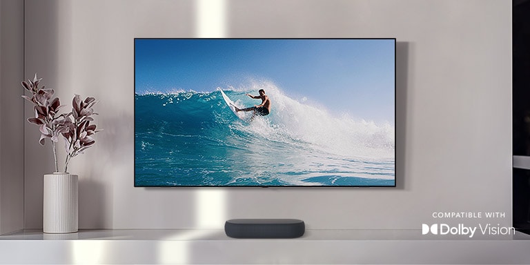 TV is on the wall. TV shows a big, swirling wave hitting the entire city. LG Eclair is right below TV on a white shelf with a sub-woofer right next. Dolby Atmos and DTS X logo shown on middle bottom of image.