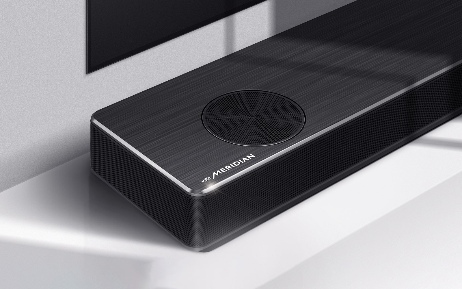Крупный план левой стороны саундбара LG с лого Meridian в левом нижнем углу. Также виден левый нижний край телевизора.